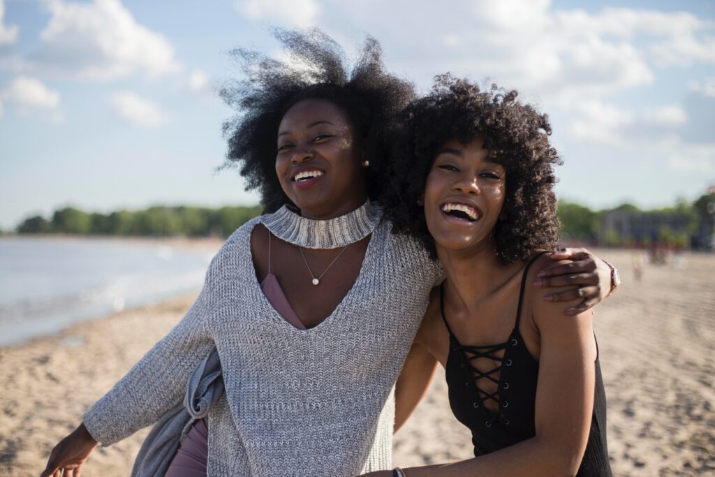 Two women laughing and embracing on a sunny beach, celebrating and enjoying each other’s company.