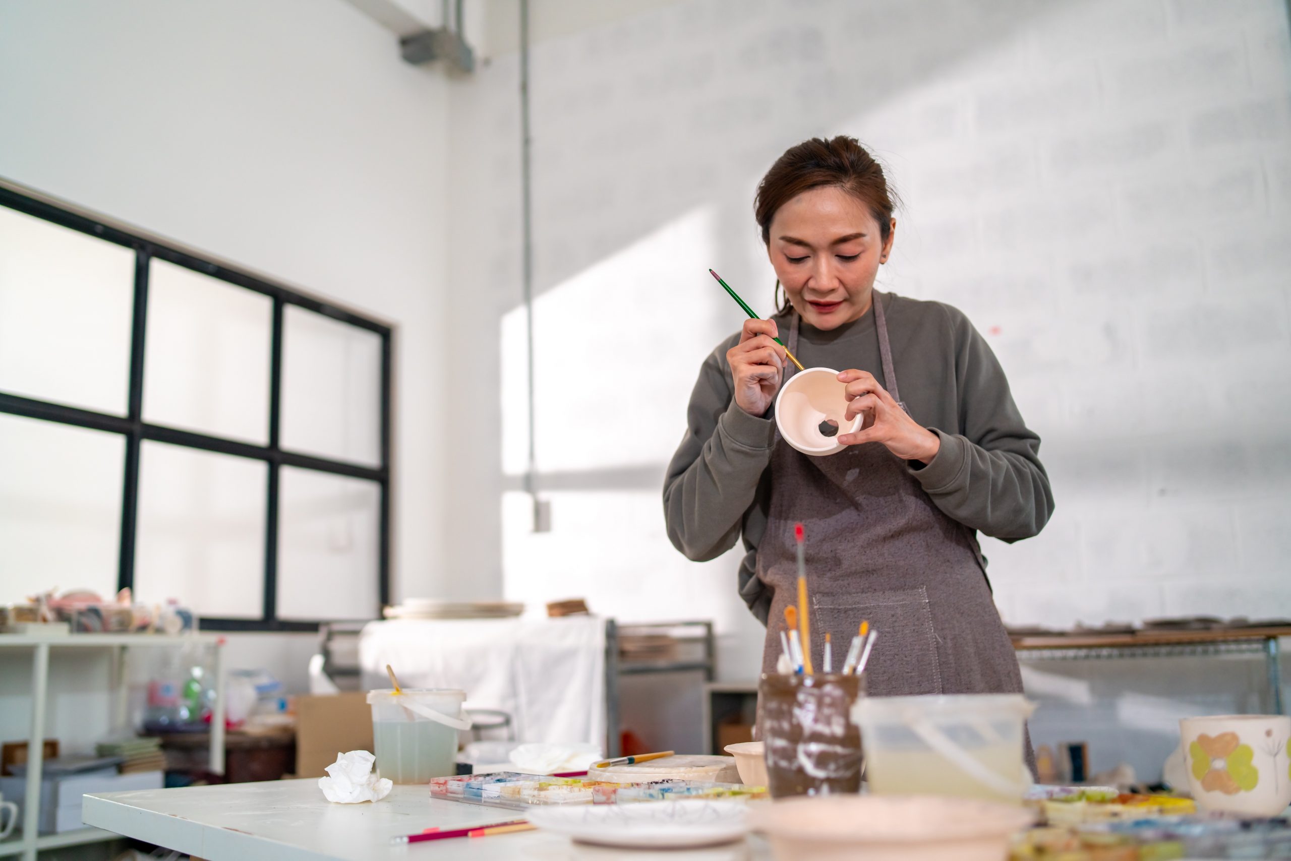Mothers-Day-Event-Ideas-Pottery-workshop-getty-images-w23YF4lrges-unsplash A woman focused on painting a ceramic piece during a pottery workshop, with brushes and art supplies spread out on the table.