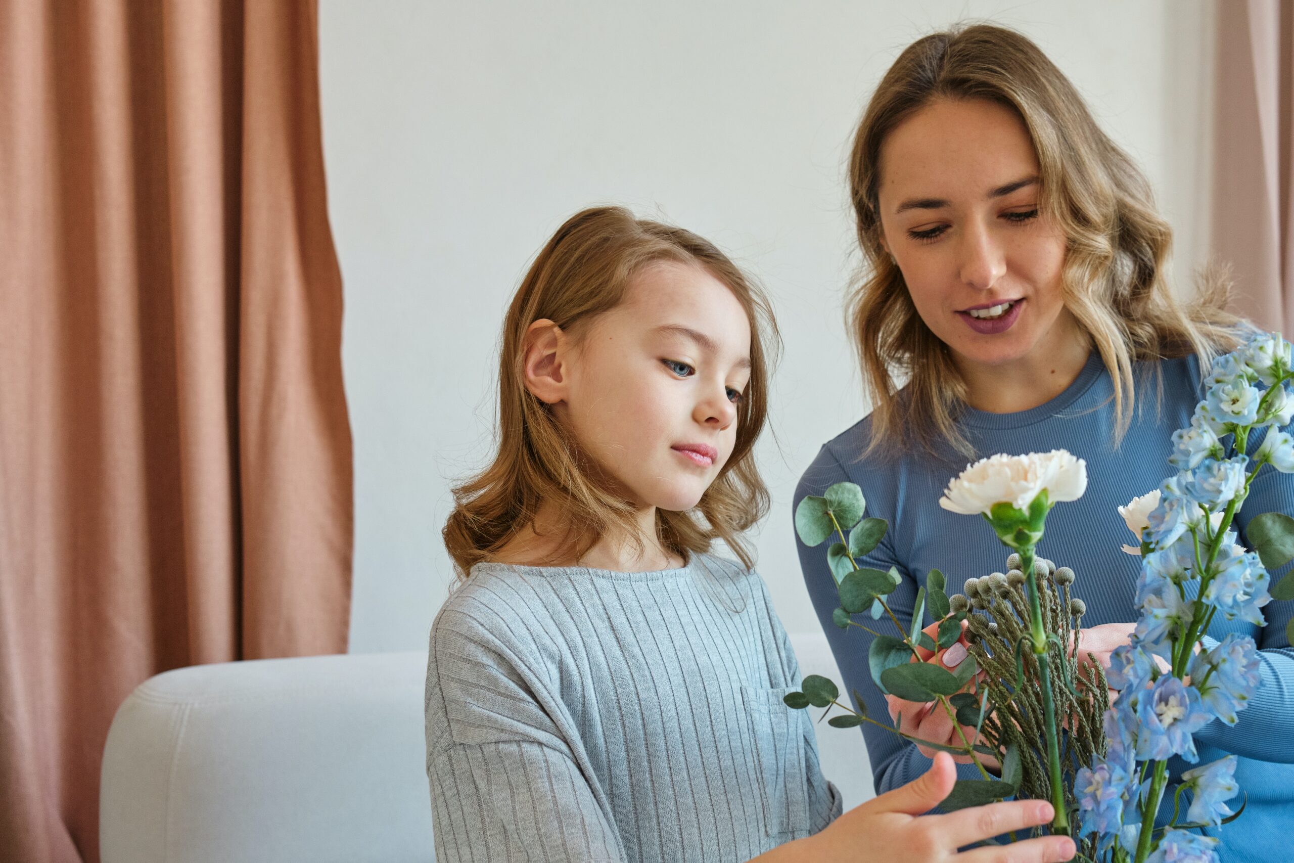 andrej-lisakov-Mothers-Day-Event-Ideas-scaled-1 A mother and young daughter smiling as they arrange a bouquet of flowers together, featuring white and blue blooms.