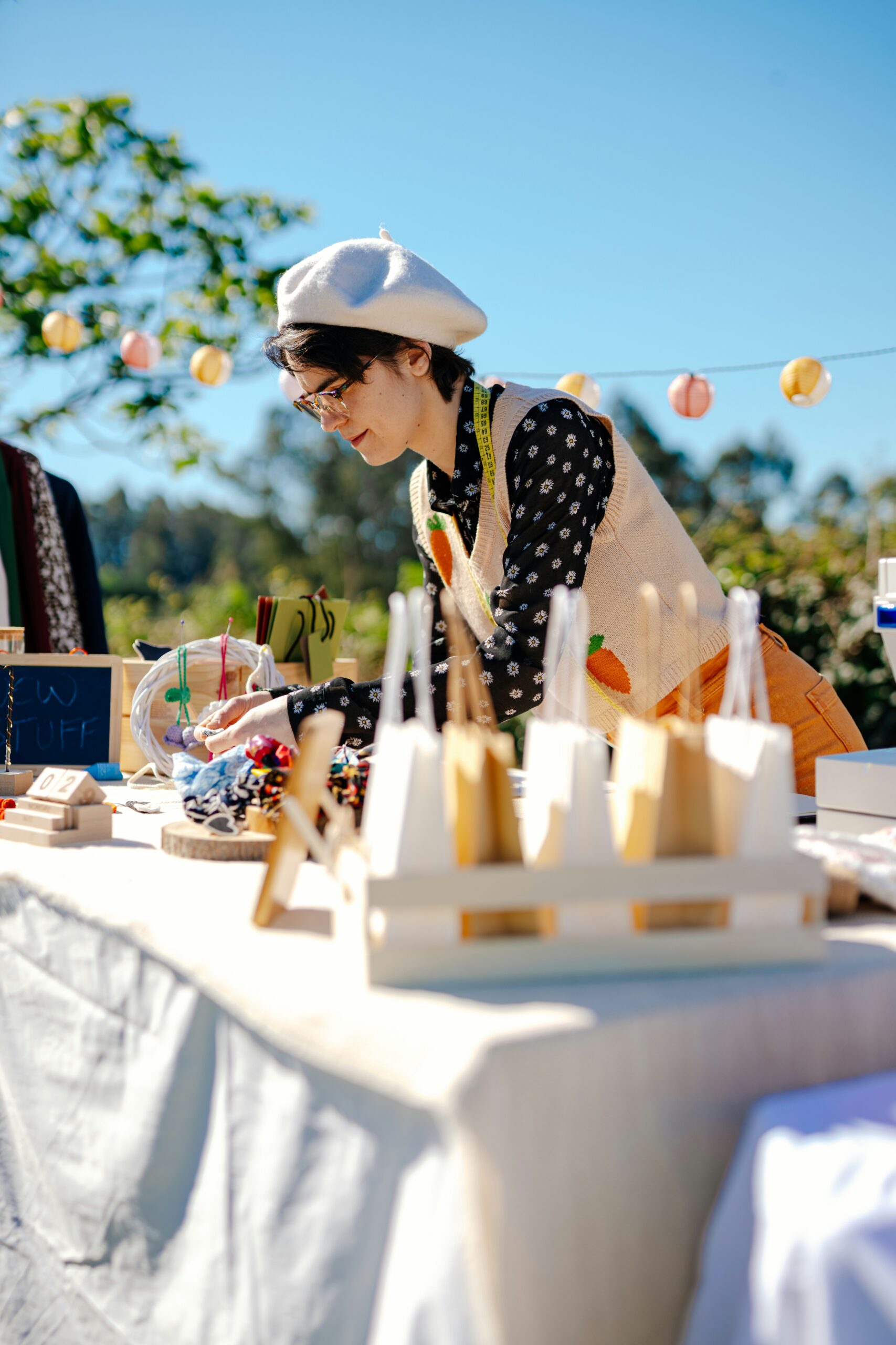 makers-market-fundraiser-vendor-booth Vendor wearing a white beret and measuring tape around their neck arranges handmade items at an outdoor makers market fundraiser booth, with colorful products and decorations displayed on the table.